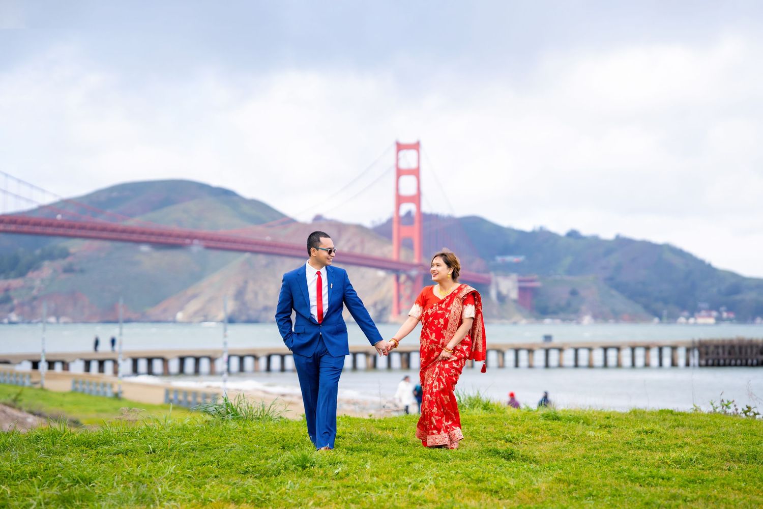 Couple in formal attire walking on grass with Golden Gate Bridge in background.