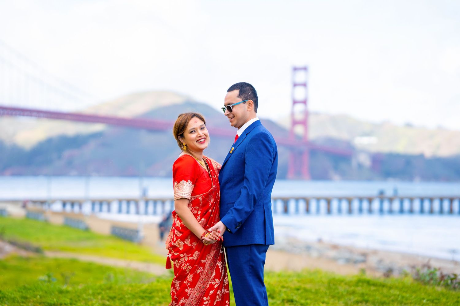 A romantic pair in red and blue formal wear stand together by the Golden Gate Bridge.