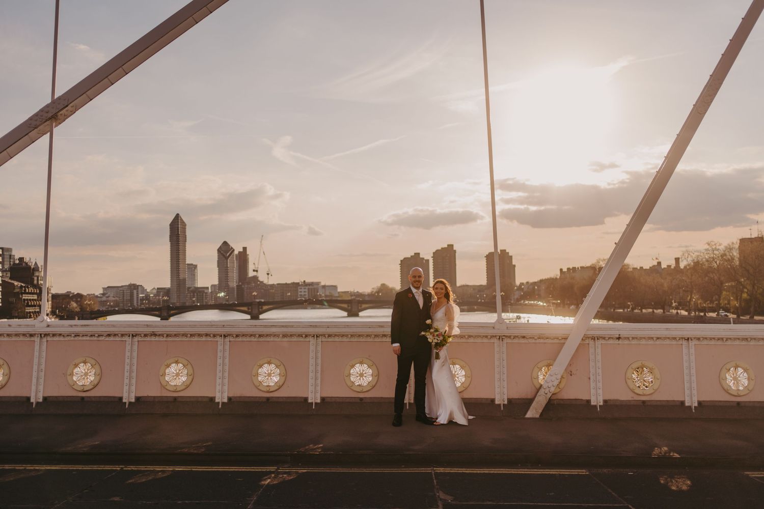 Couple on bridge at sunset with city skyline and river in background.