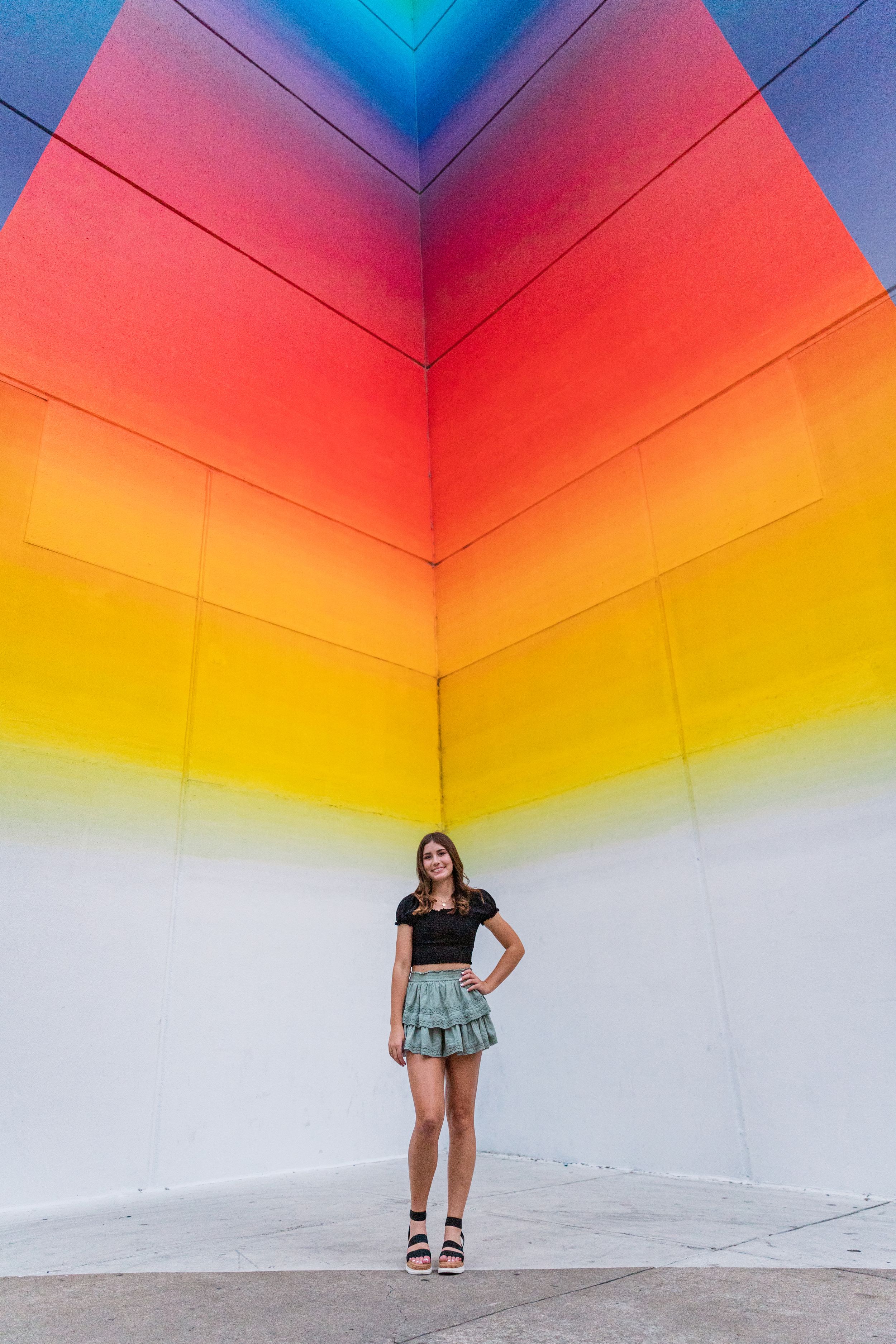 Woman poses against vibrant rainbow gradient wall mural in artistic outdoor setting.