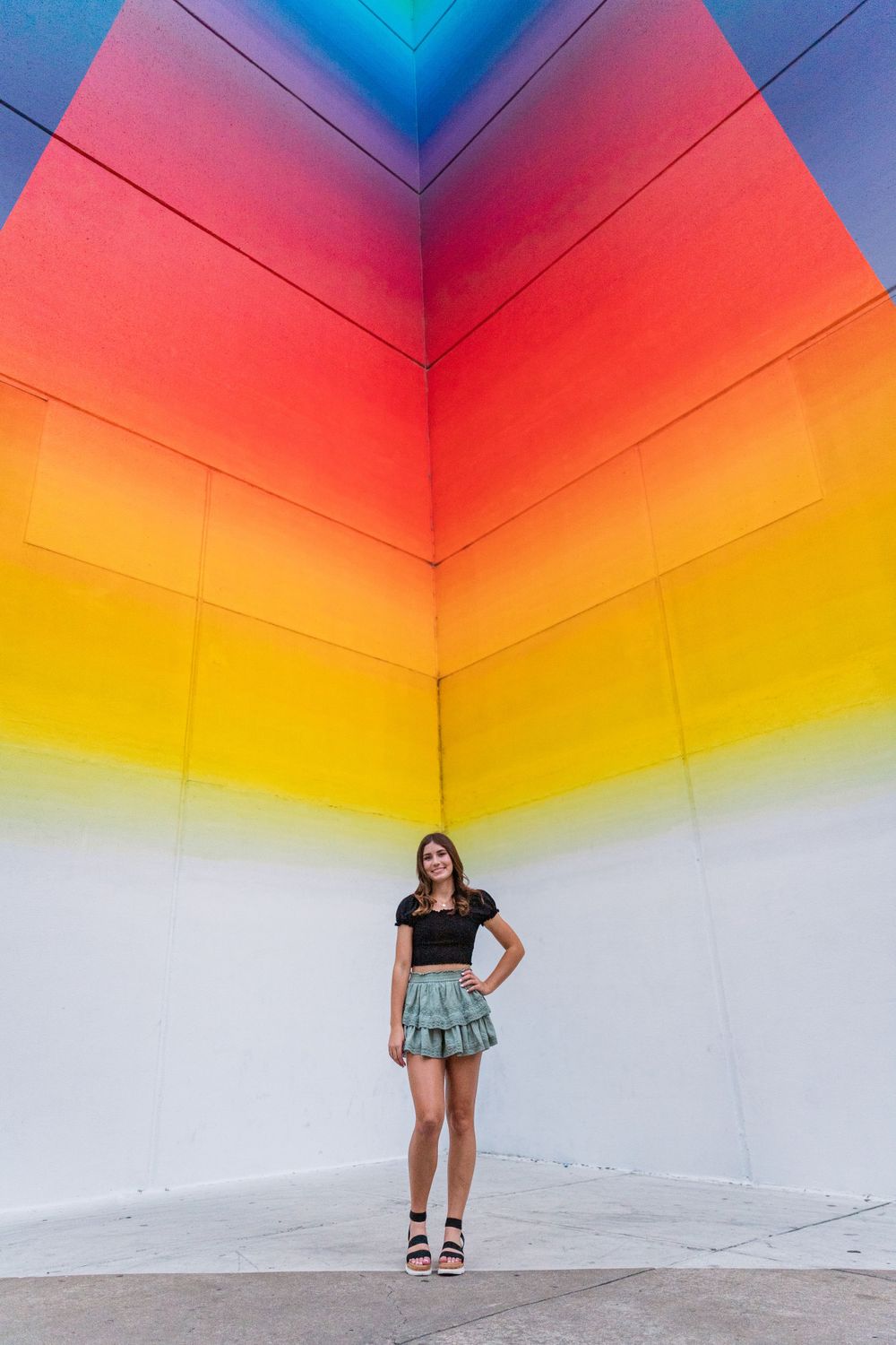 Woman poses against vibrant rainbow gradient wall mural in artistic outdoor setting.