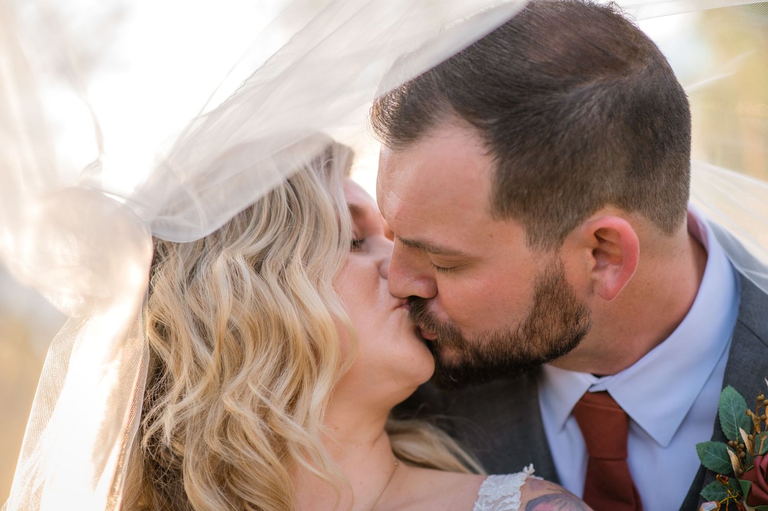 Romantic wedding couple sharing a kiss under flowing veil in golden sunlight.