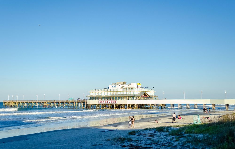 Beachfront pier with buildings and waves rolling onto sandy shore on a sunny day.
