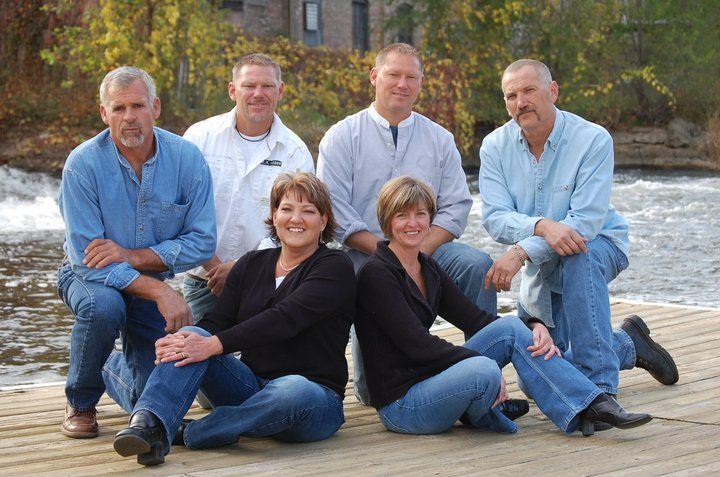 Family group photo of six people in casual blue clothing posed together on a wooden dock by the water.