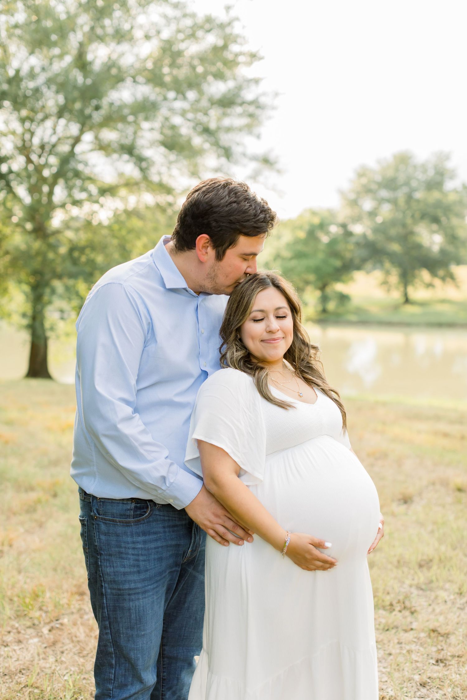 An intimate maternity portrait of a couple embracing in a sunlit field near a pond.