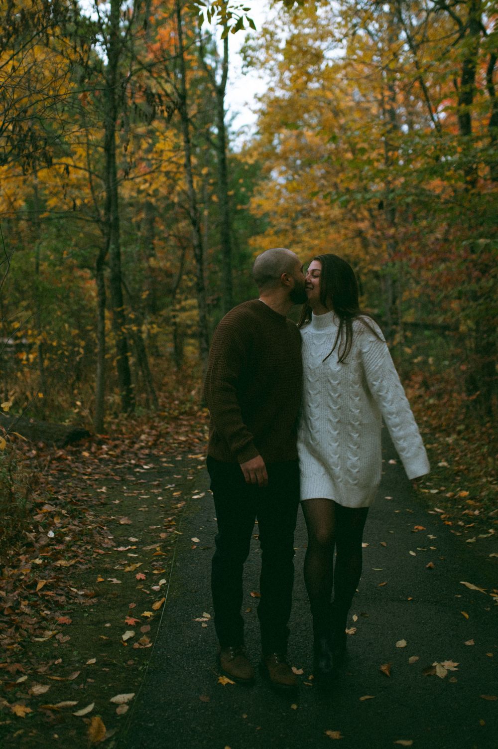 Two figures share an intimate moment on a leaf-covered trail in autumn.