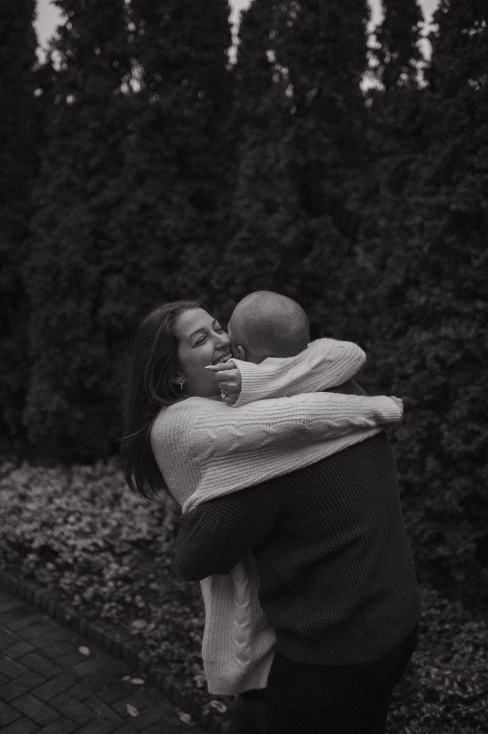 A couple embraces joyfully outdoors in a black and white photo with trees in the background.