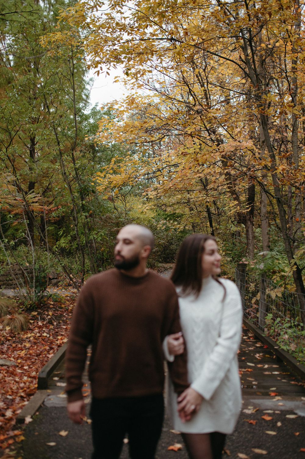 A couple walking hand in hand on a forest path covered in autumn leaves.