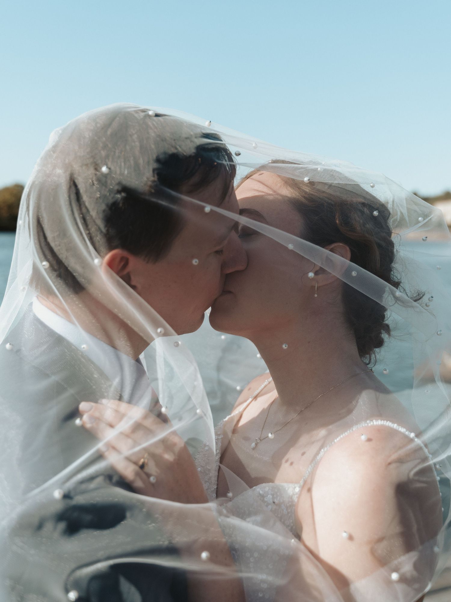 Close up photo of a newlywed couple kissing underneath a veil speckled with pearls.