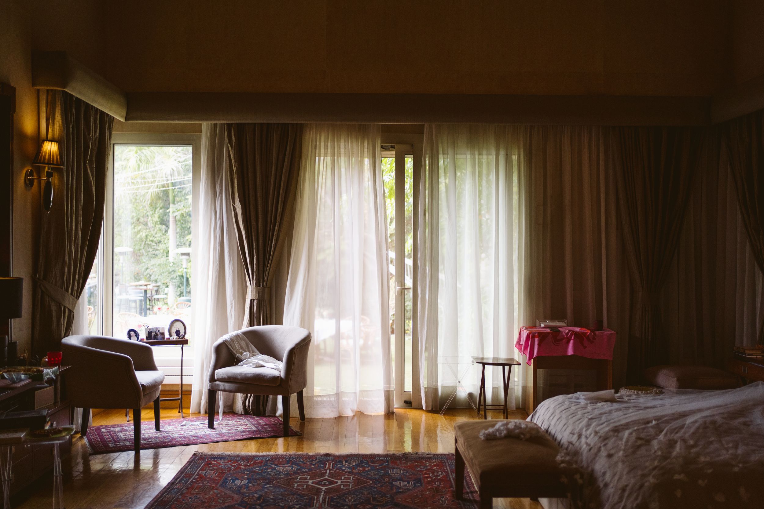 Sunlit room interior with sheer curtains, chairs and bed visible.