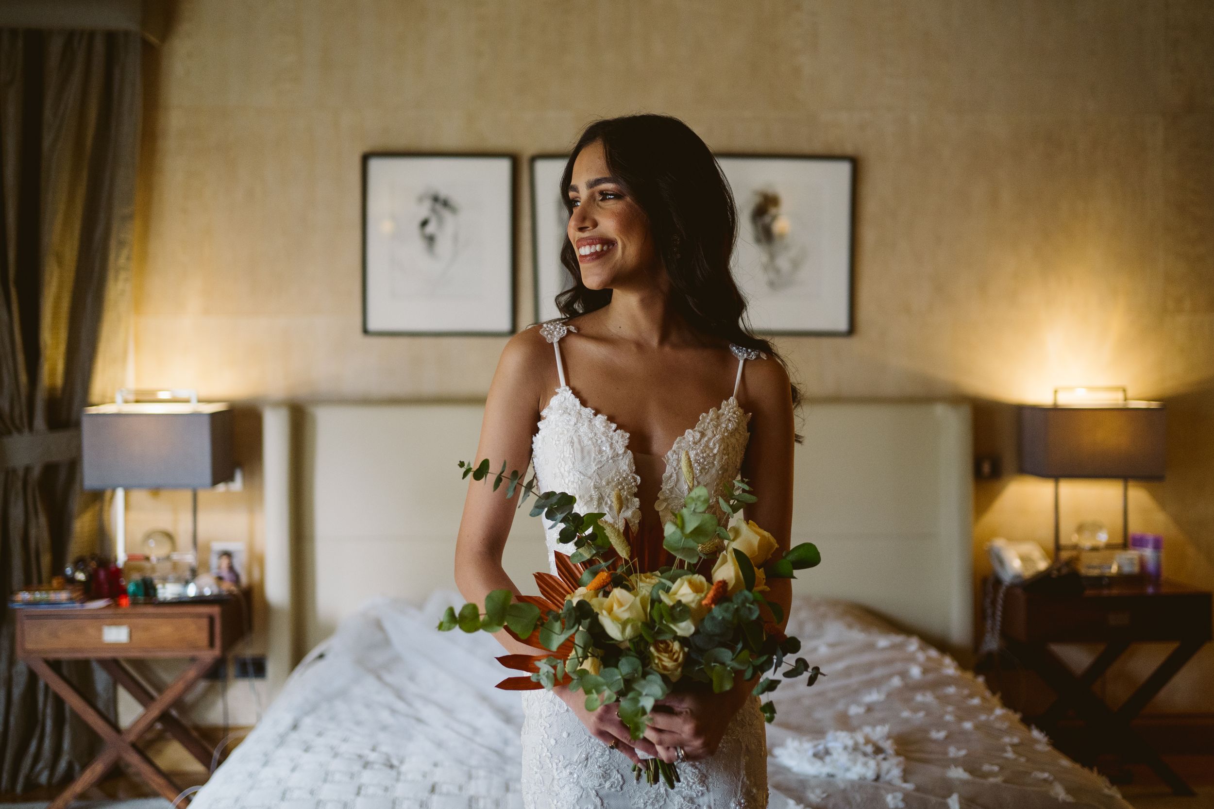 Bride holding colorful bouquet while laughing in elegant bedroom setting.