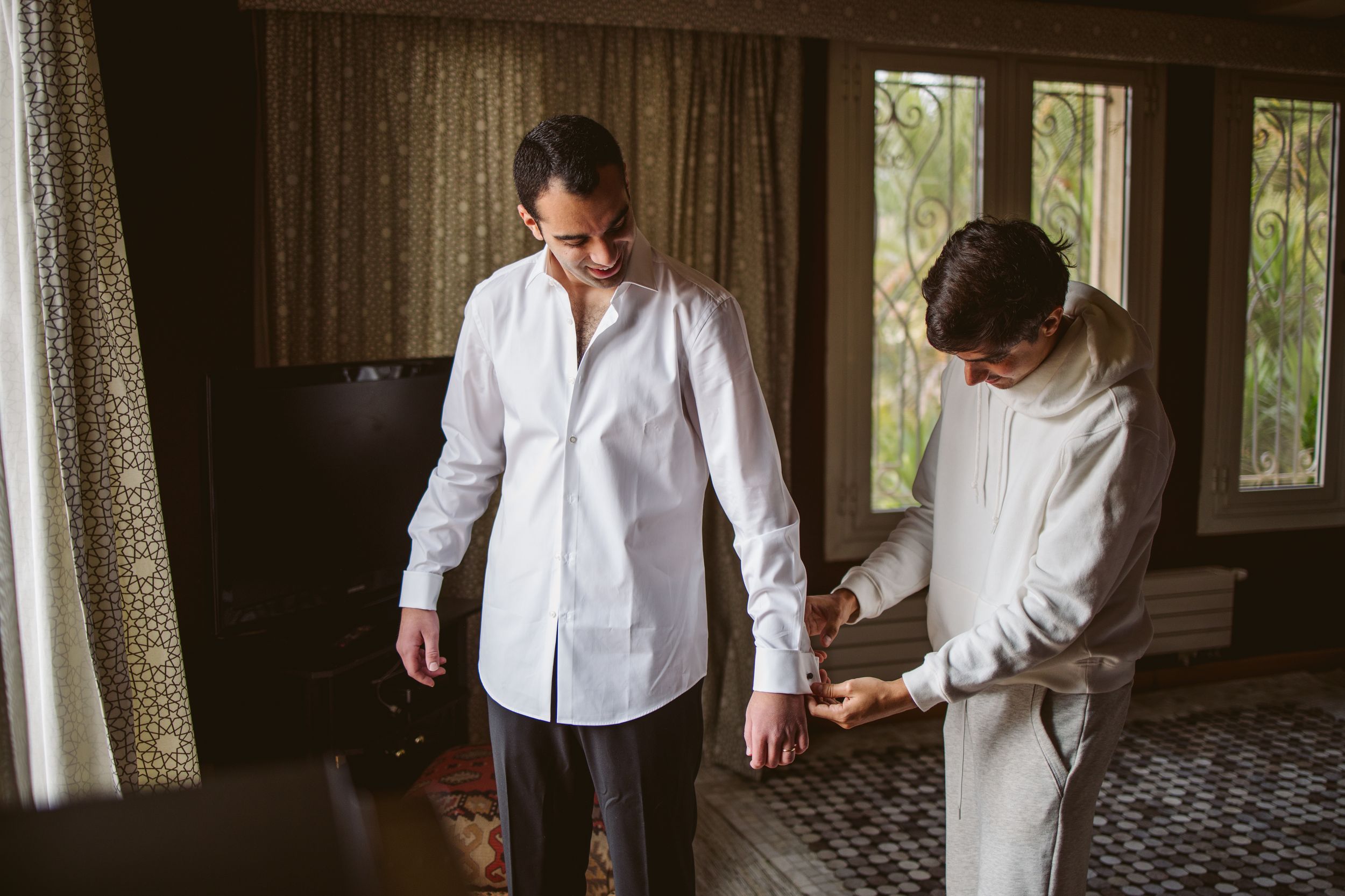 Two people in white shirts getting ready in a sunlit room with curtained windows.