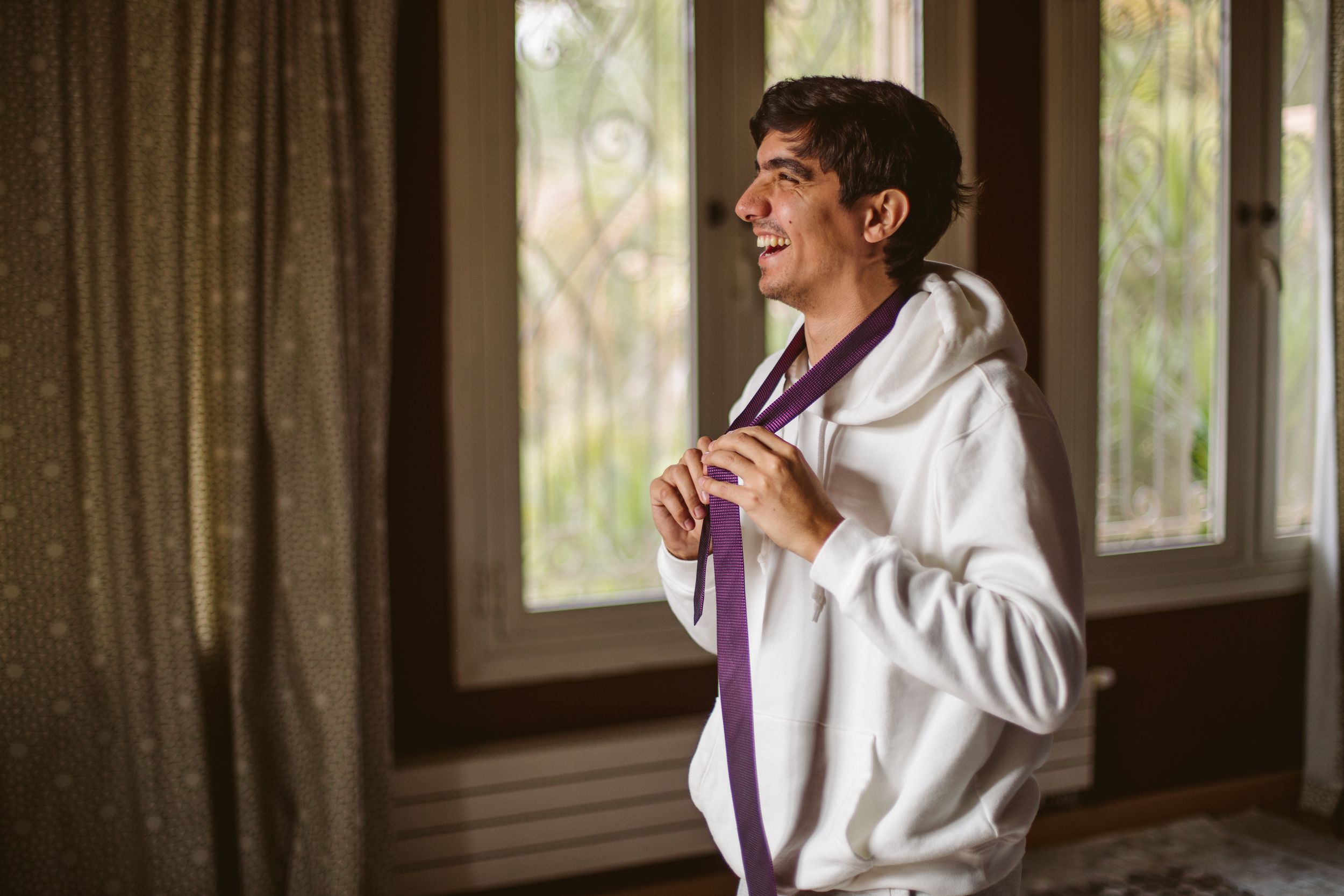 Person in white adjusting purple tie while getting dressed near window.
