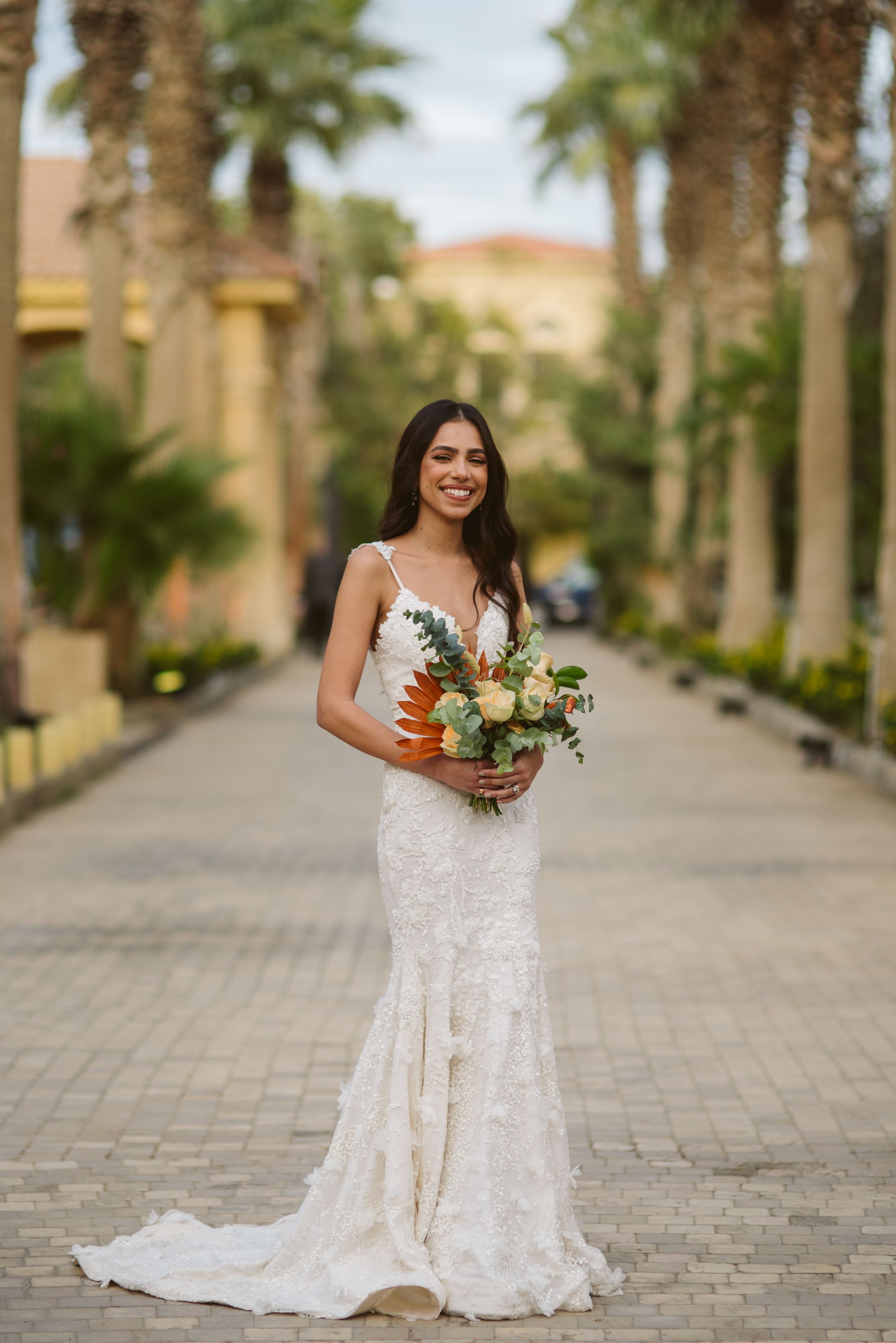 Bride in lace mermaid gown holds tropical bouquet on palm-lined pathway.