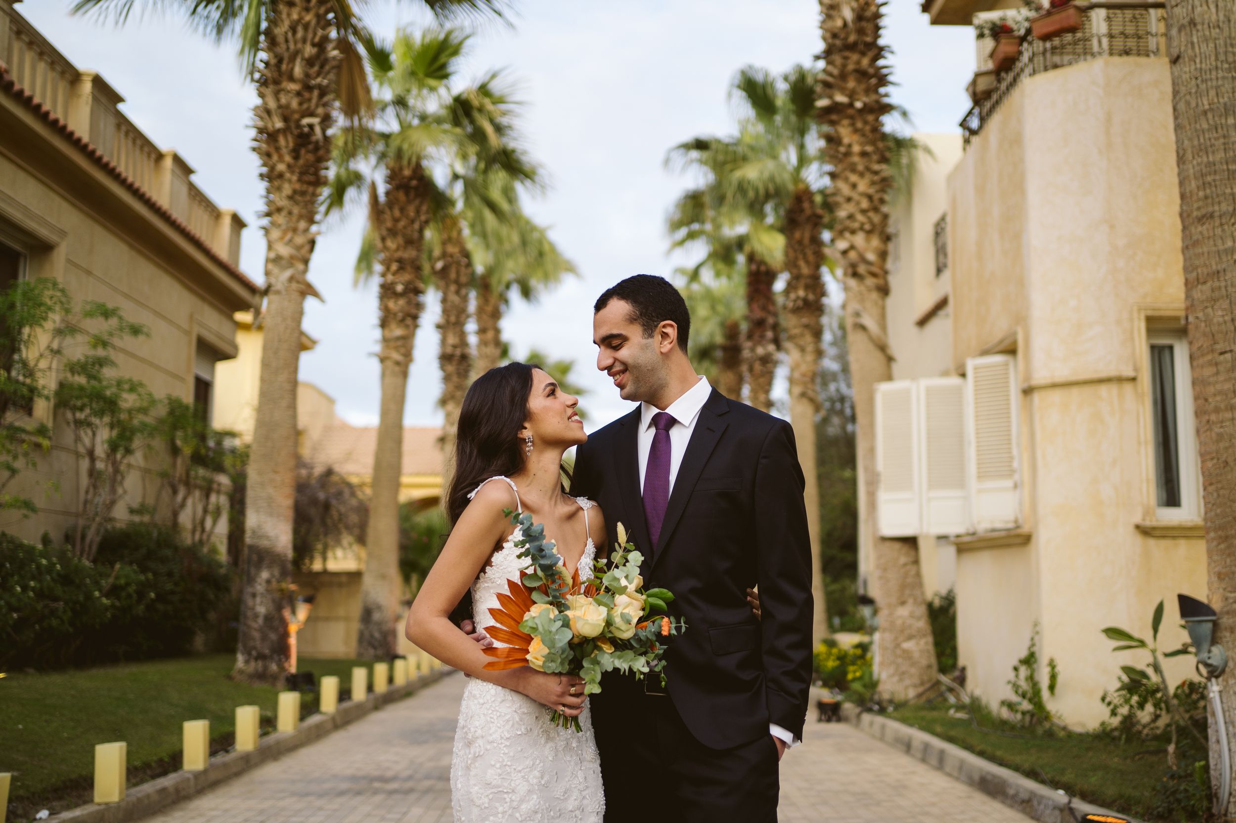 Newlyweds share loving glance while walking along palm-lined resort pathway.