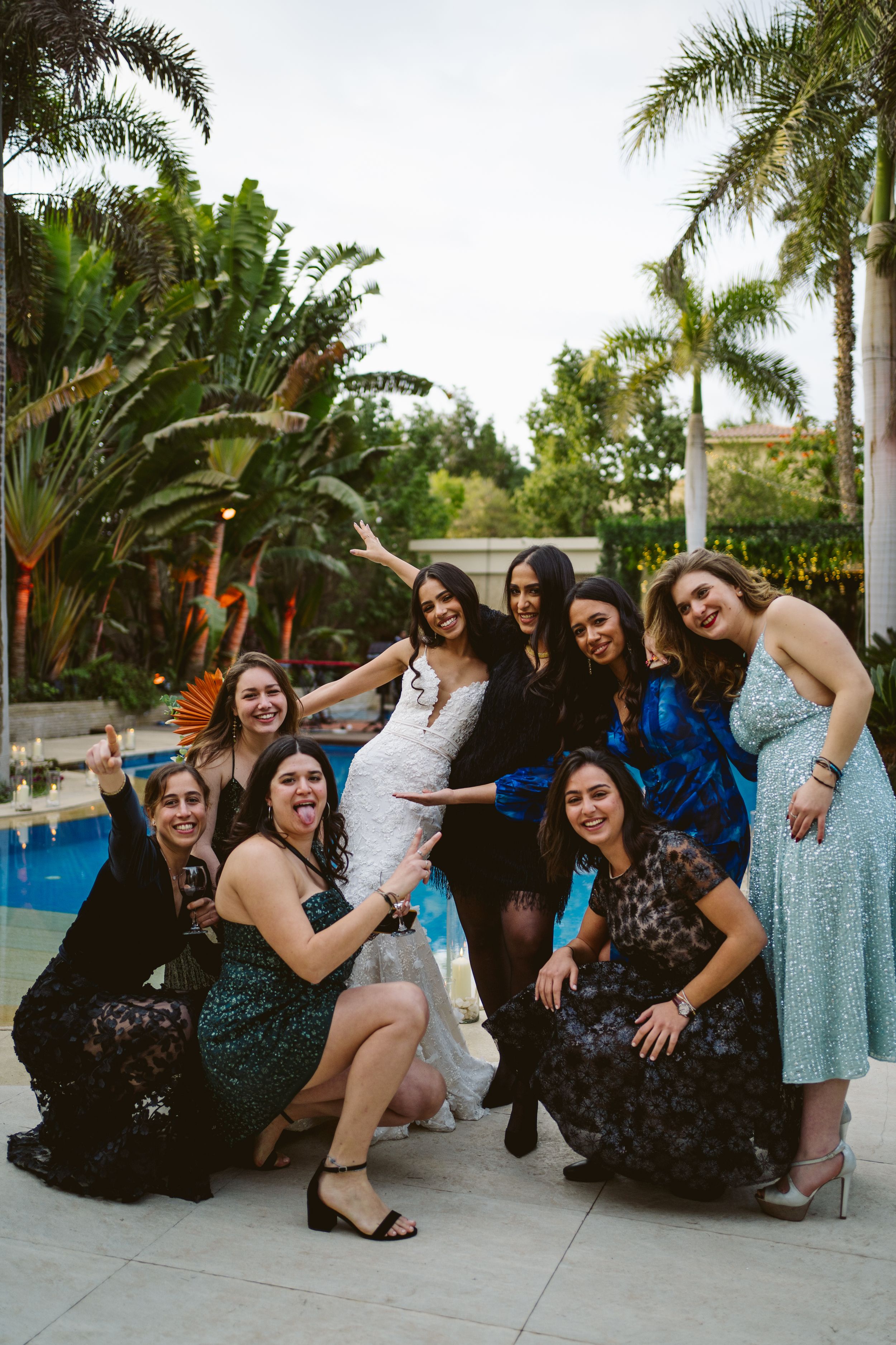 Bridesmaids in formal dresses pose together by a pool during an outdoor wedding celebration.