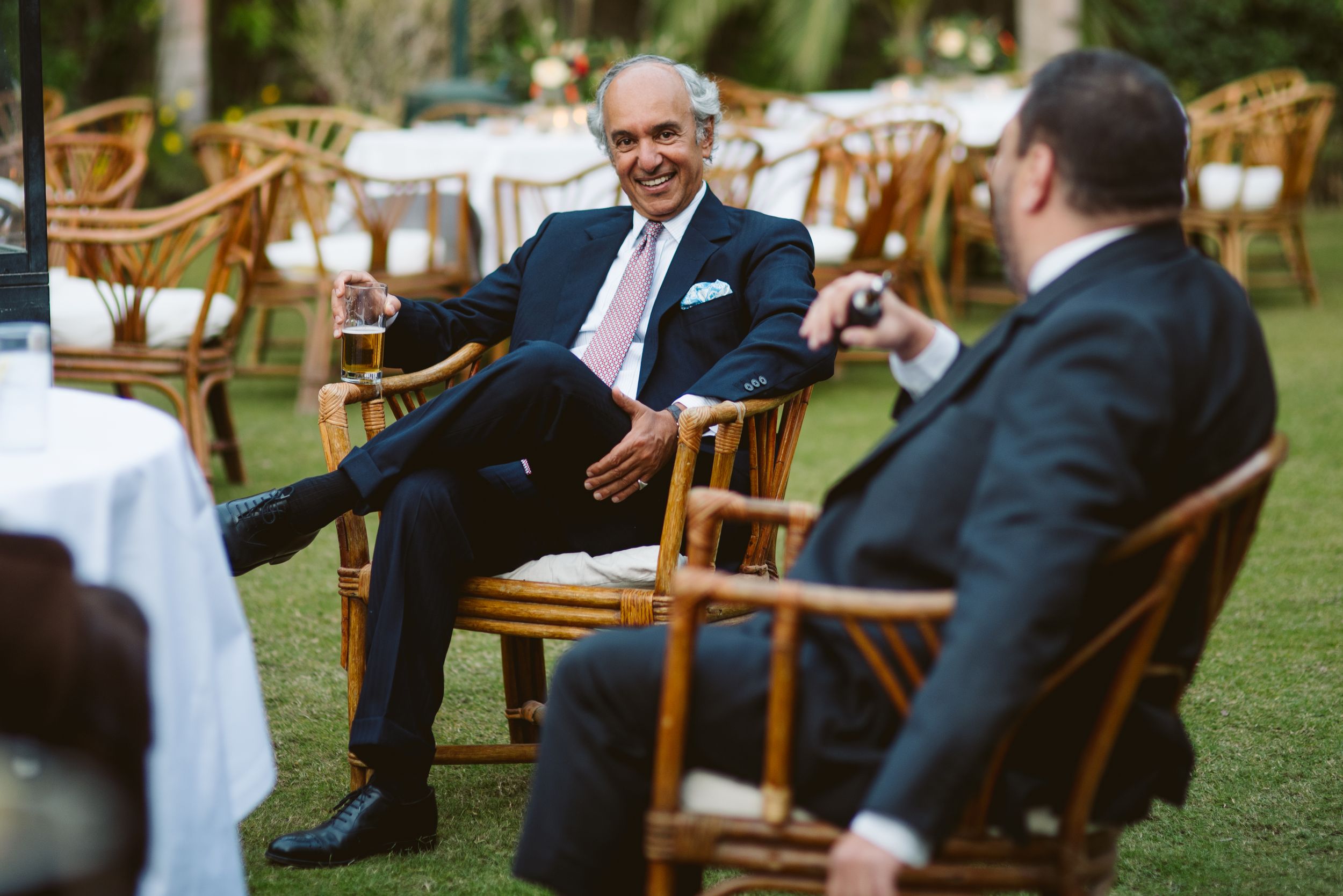 Two men in suits having a conversation while seated in wooden chairs on a lawn.