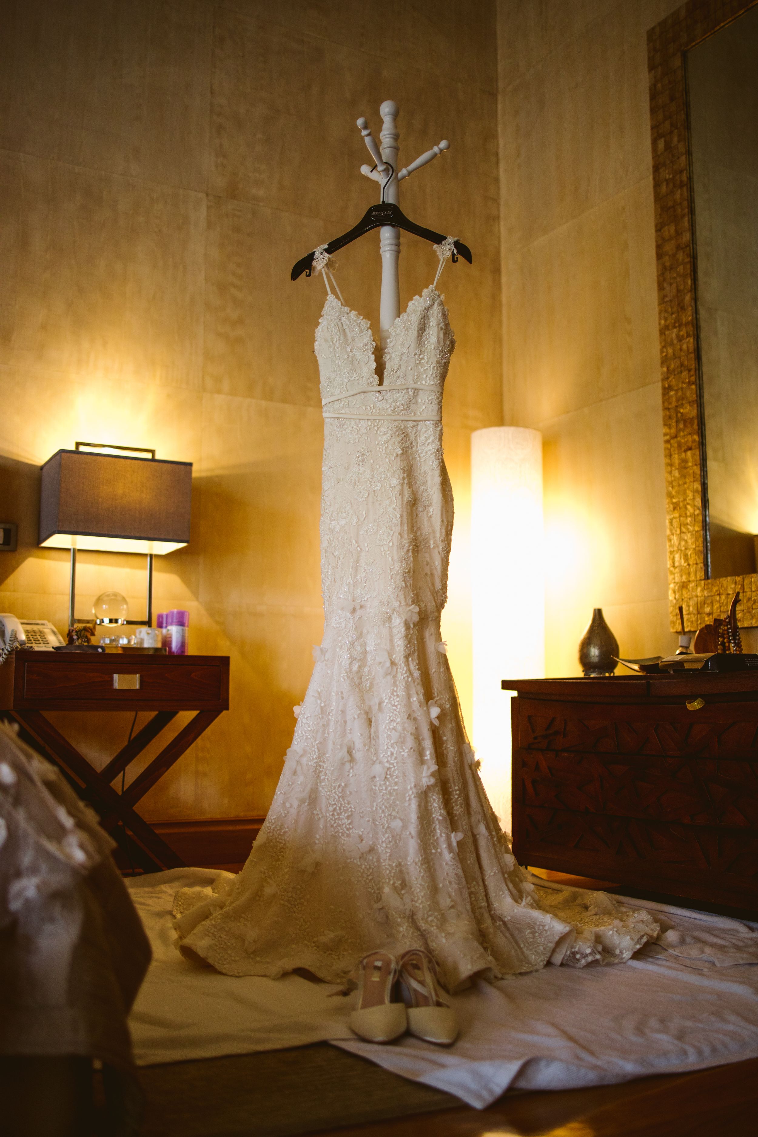 Elegant lace wedding gown displayed on a hanger in a golden-lit room with furniture.