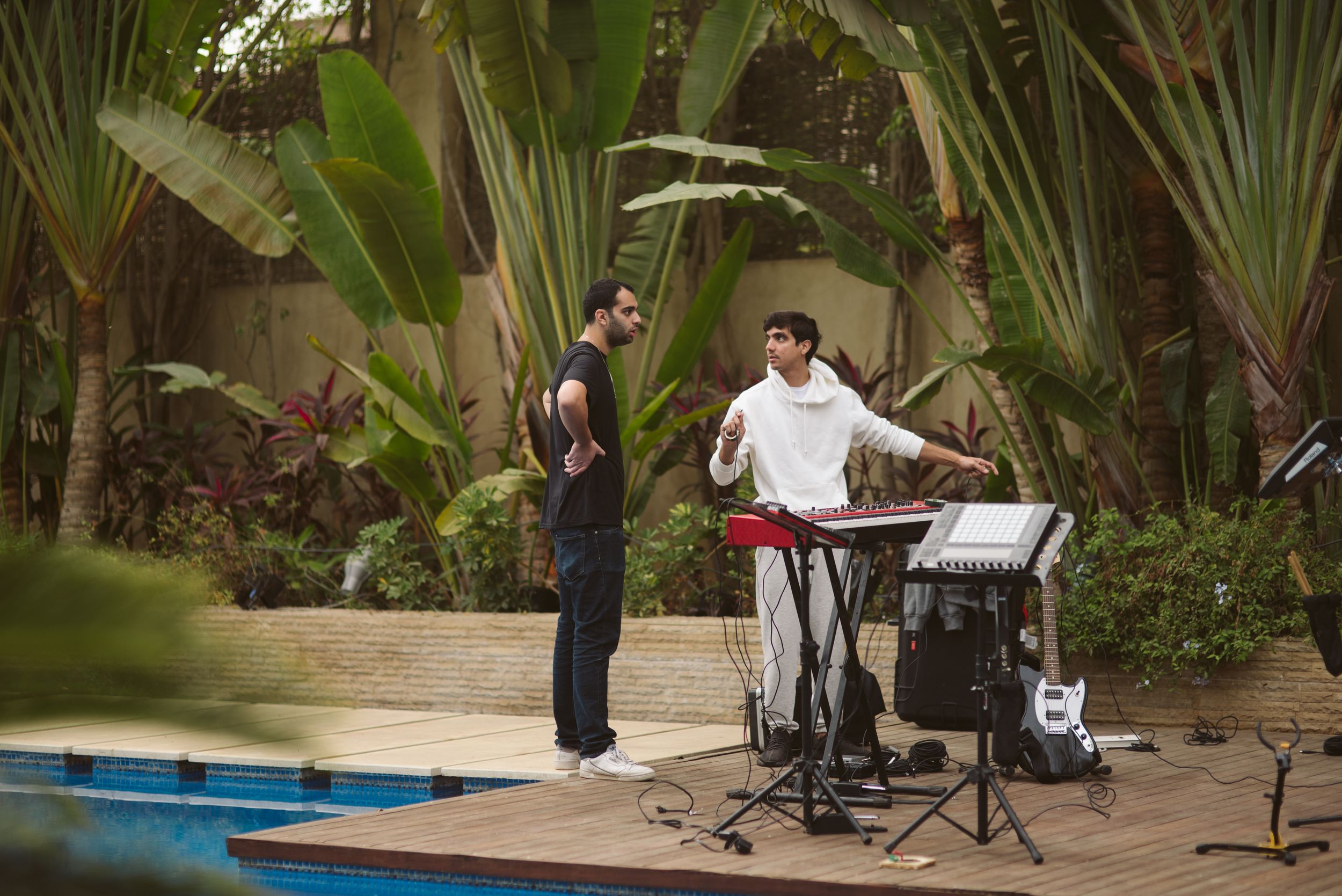 Musicians perform poolside with instruments and equipment set up in a tropical garden setting.