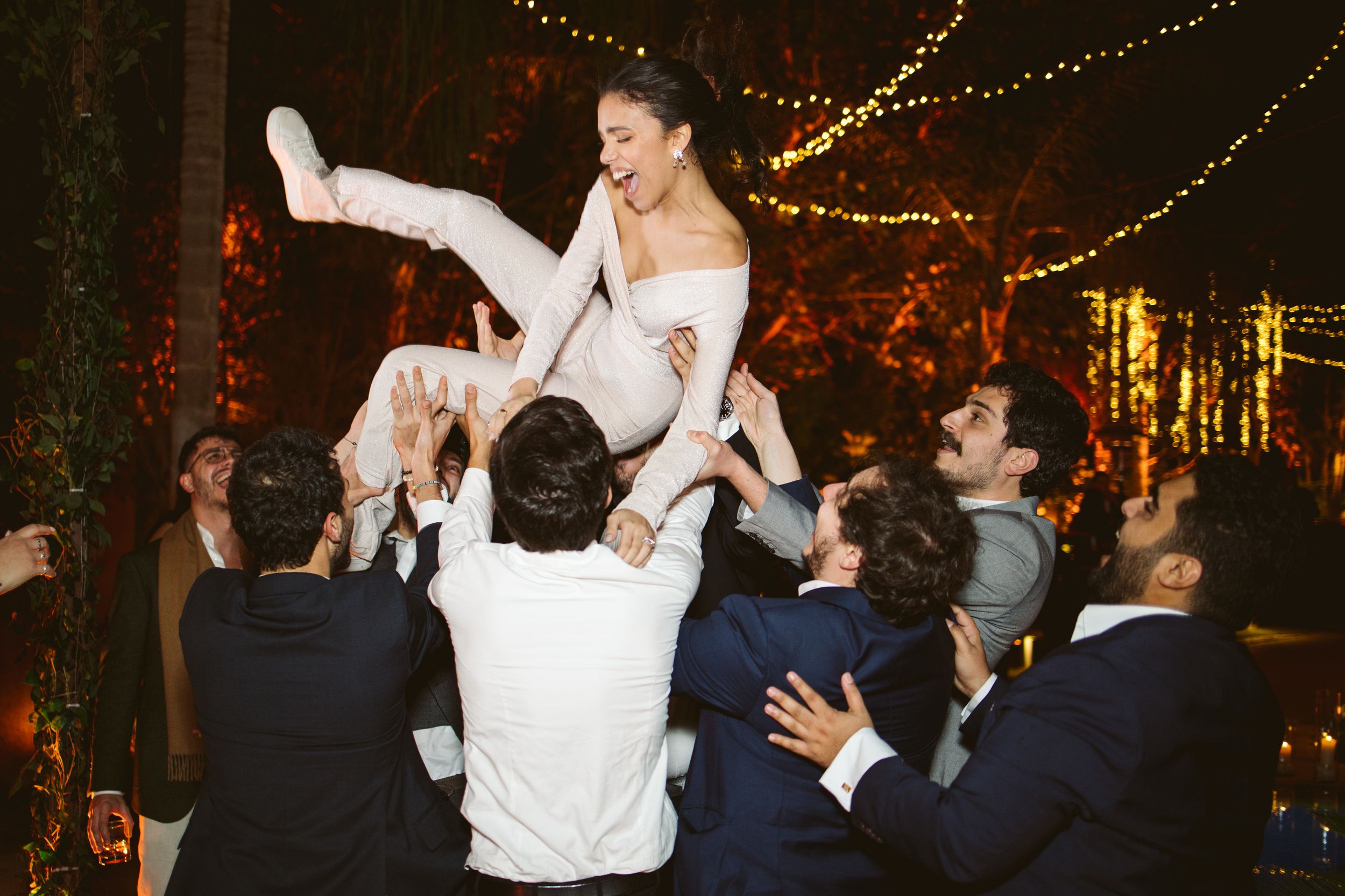 Wedding celebration with guests lifting the bride during a lively dance party with twinkle lights.
