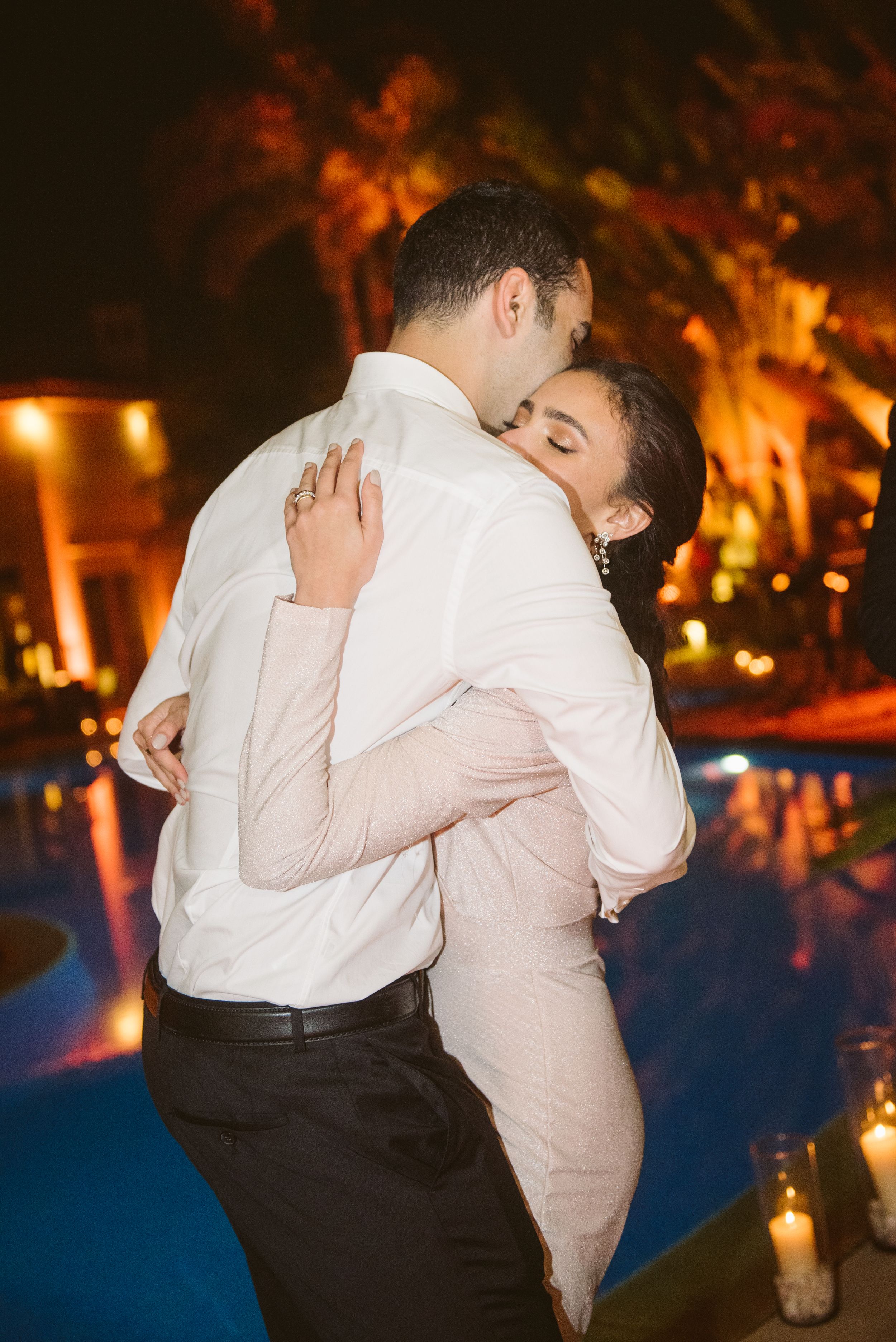 Intimate slow dance moment between newlyweds beside illuminated pool at night.