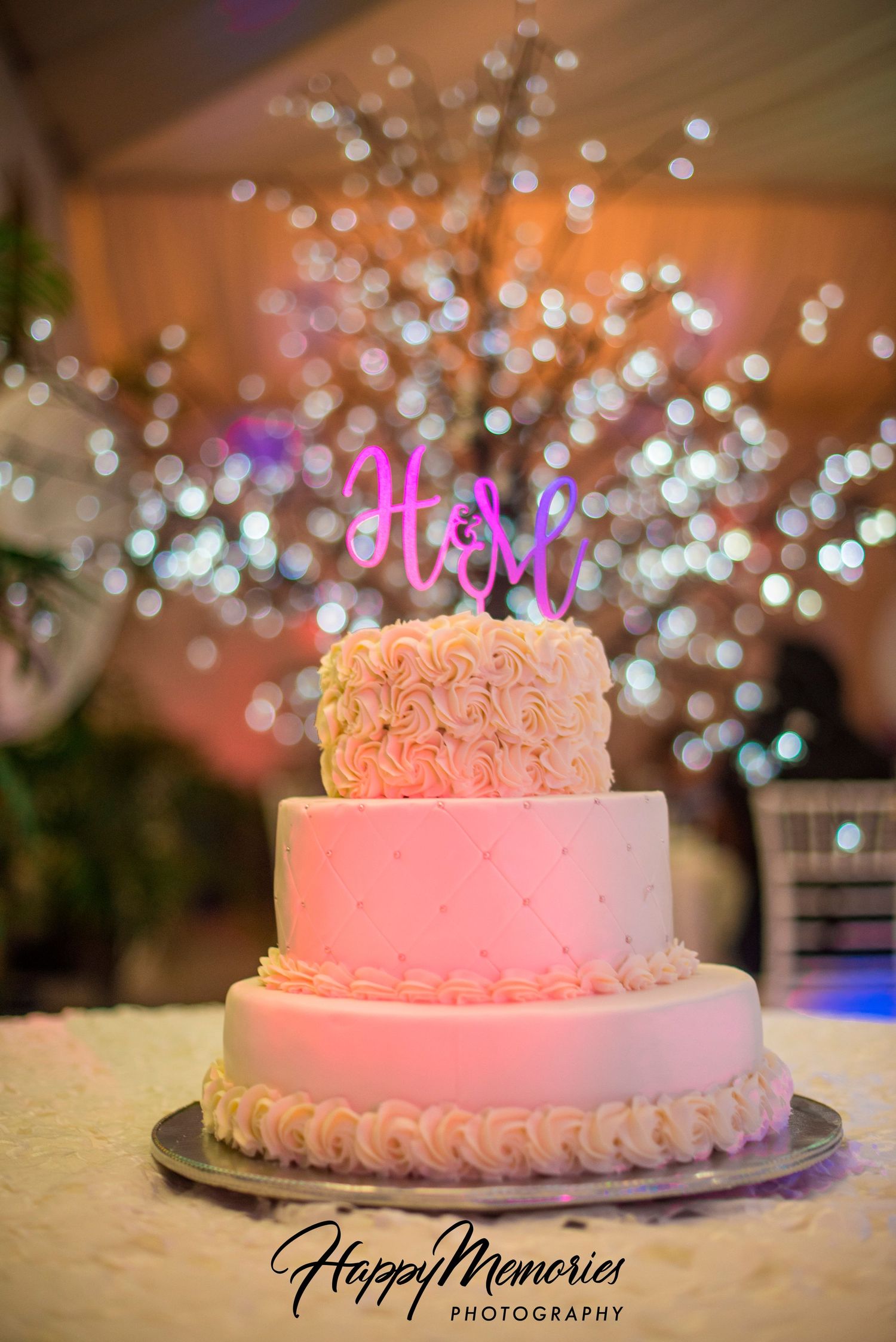 Elegant three-tier wedding cake with pink roses and twinkle light bokeh background.