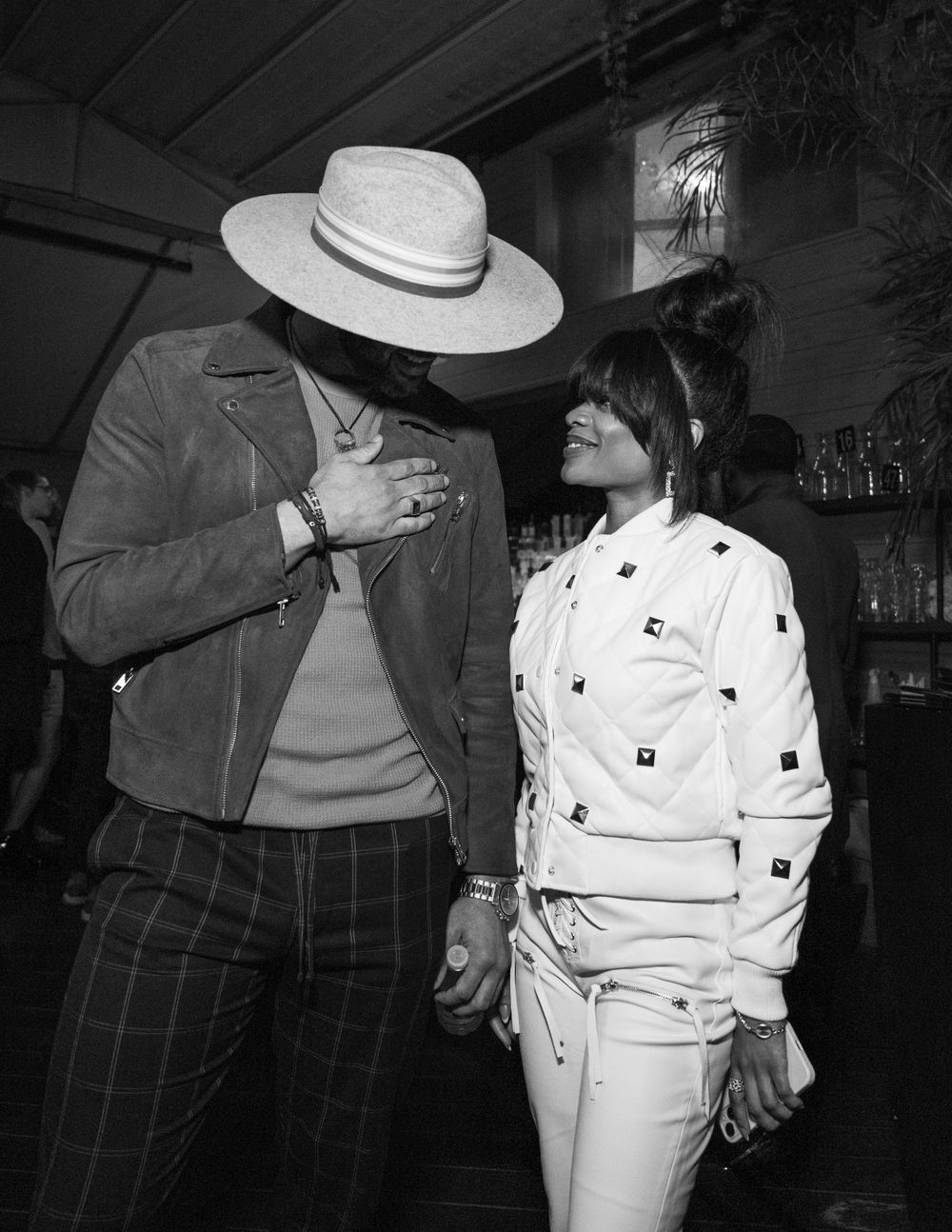 Person wearing a white patterned outfit stands next to someone in a hat in a casual indoor setting.