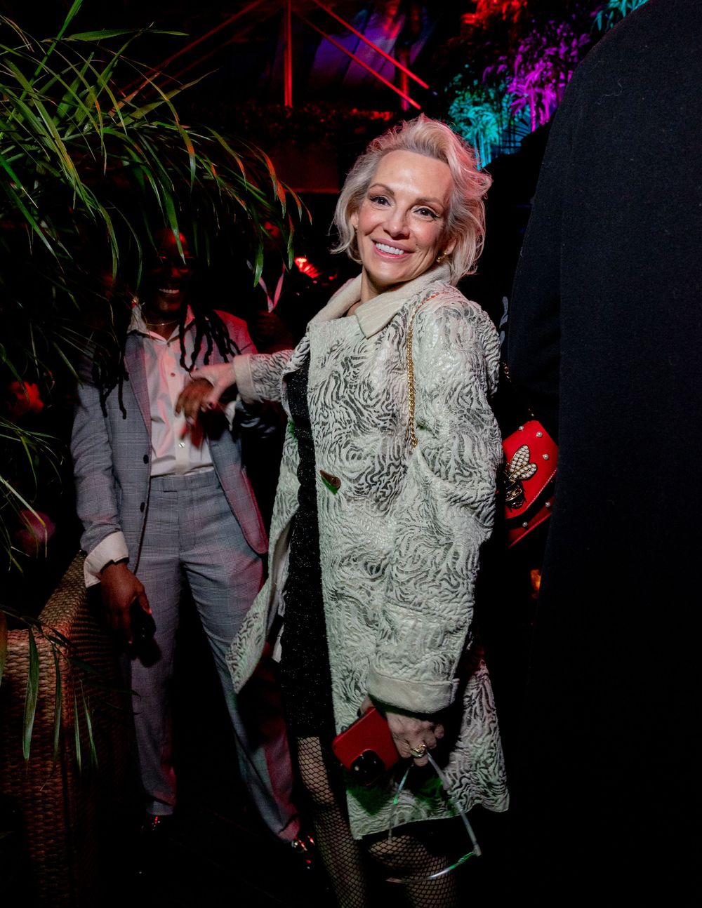 Party scene with someone wearing a light patterned coat amidst colorful lighting and tropical decor.