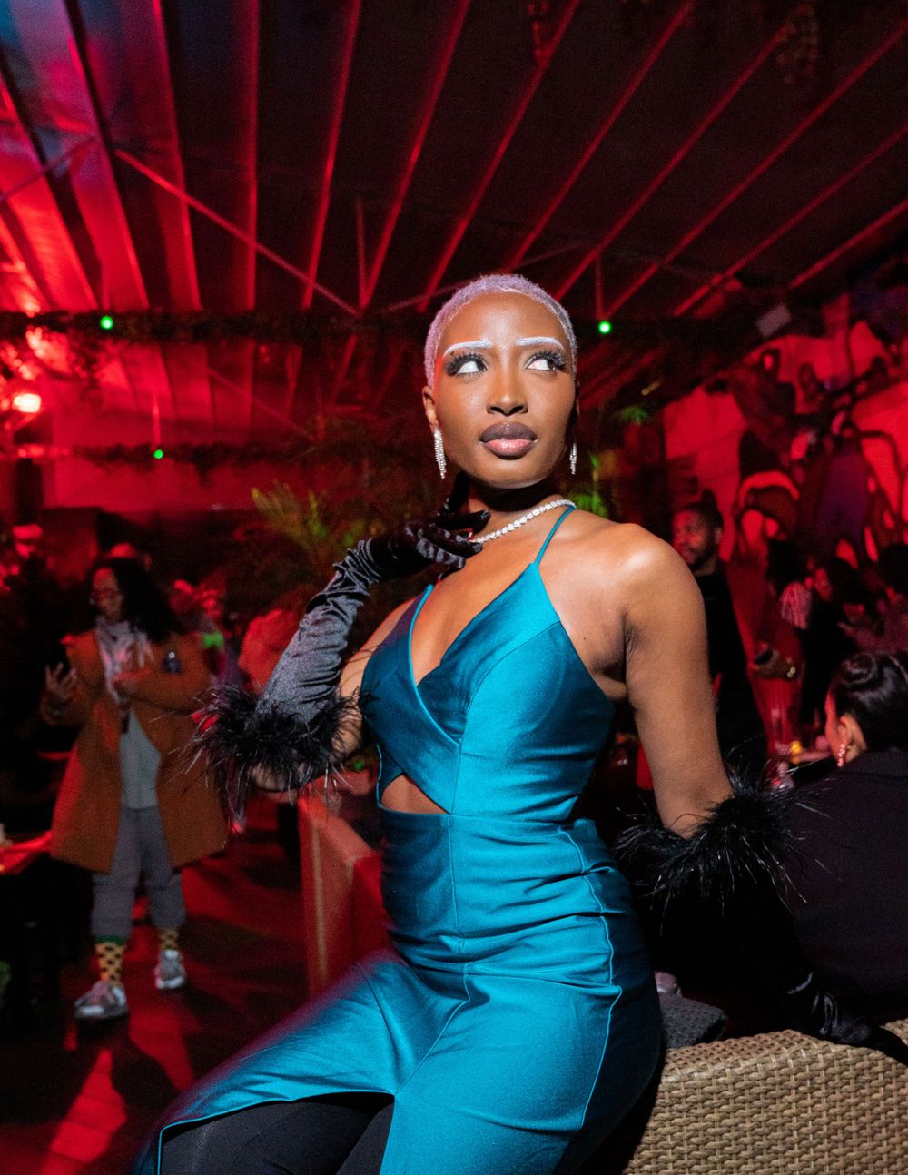 Performer in bright blue jumpsuit and black feathered gloves strikes a pose under red lighting.