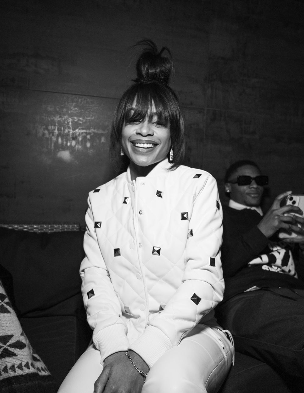 Black and white photo showing someone in a white patterned outfit with a top knot hairstyle smiling warmly.
