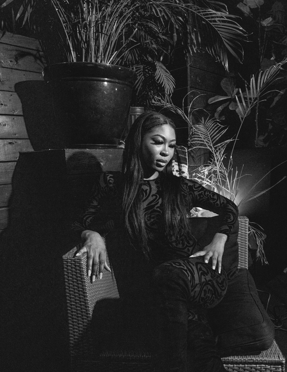 Black and white portrait showing someone reclining on furniture near potted plants.
