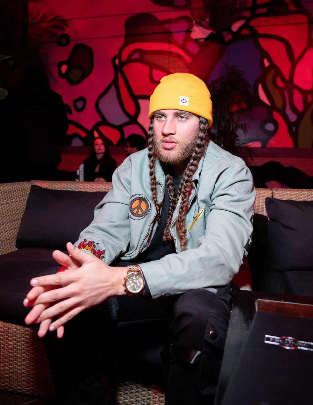 Person in yellow beanie and gray jacket sitting on couch with braided hair.