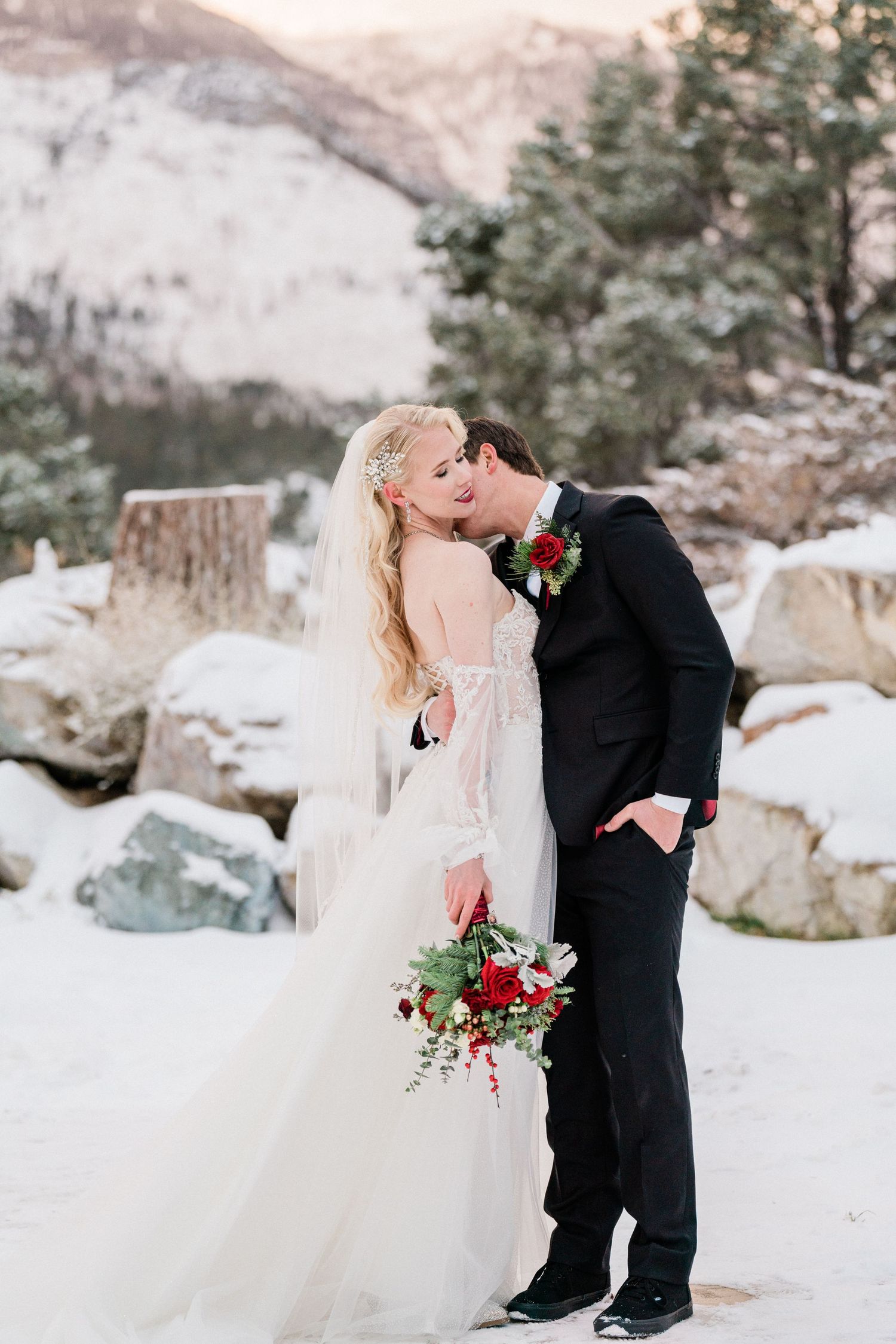 A winter wedding couple kisses in a snowy mountain landscape with red flowers.