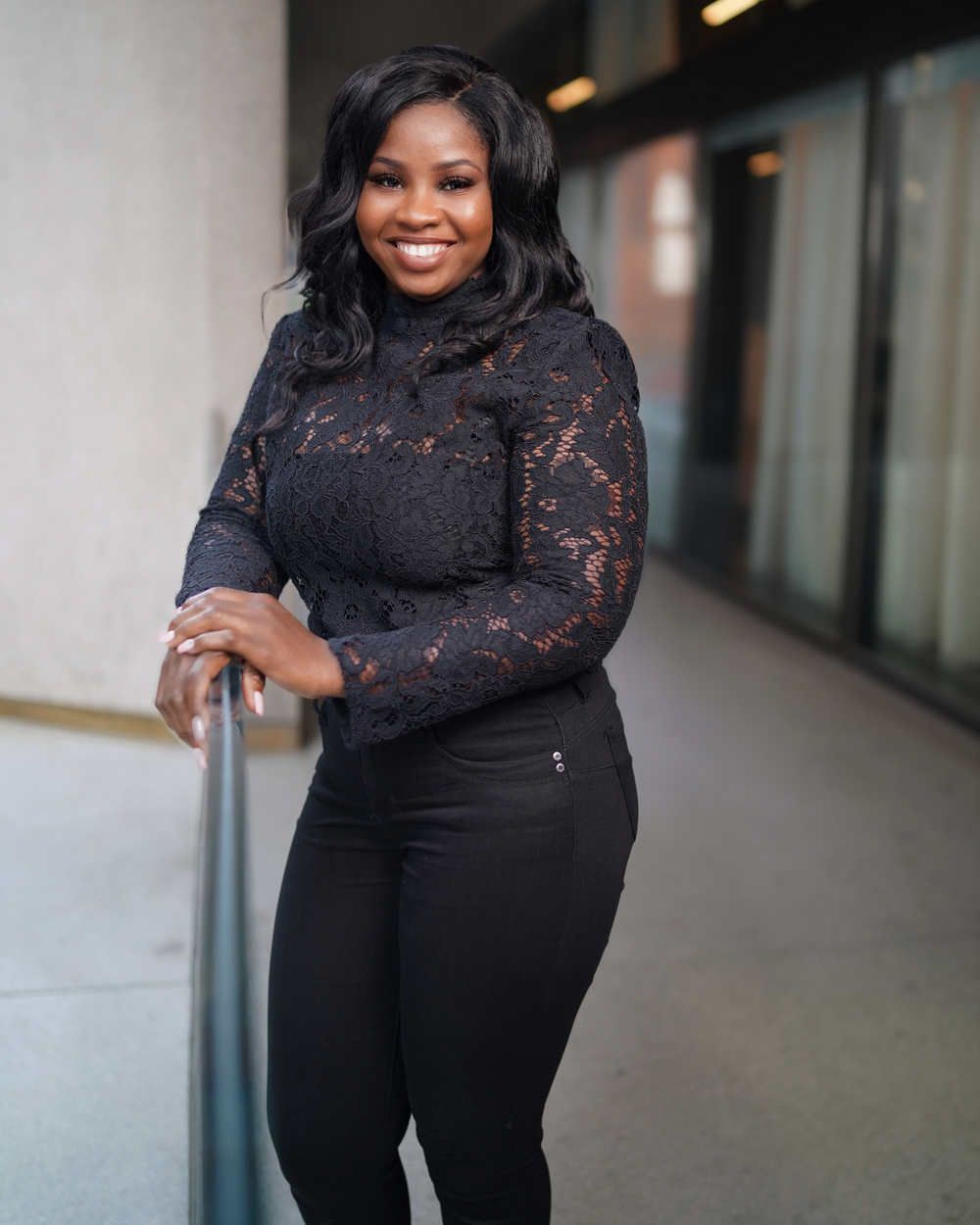 Smiling woman in black lace turtleneck top and black pants posing in a modern hallway.