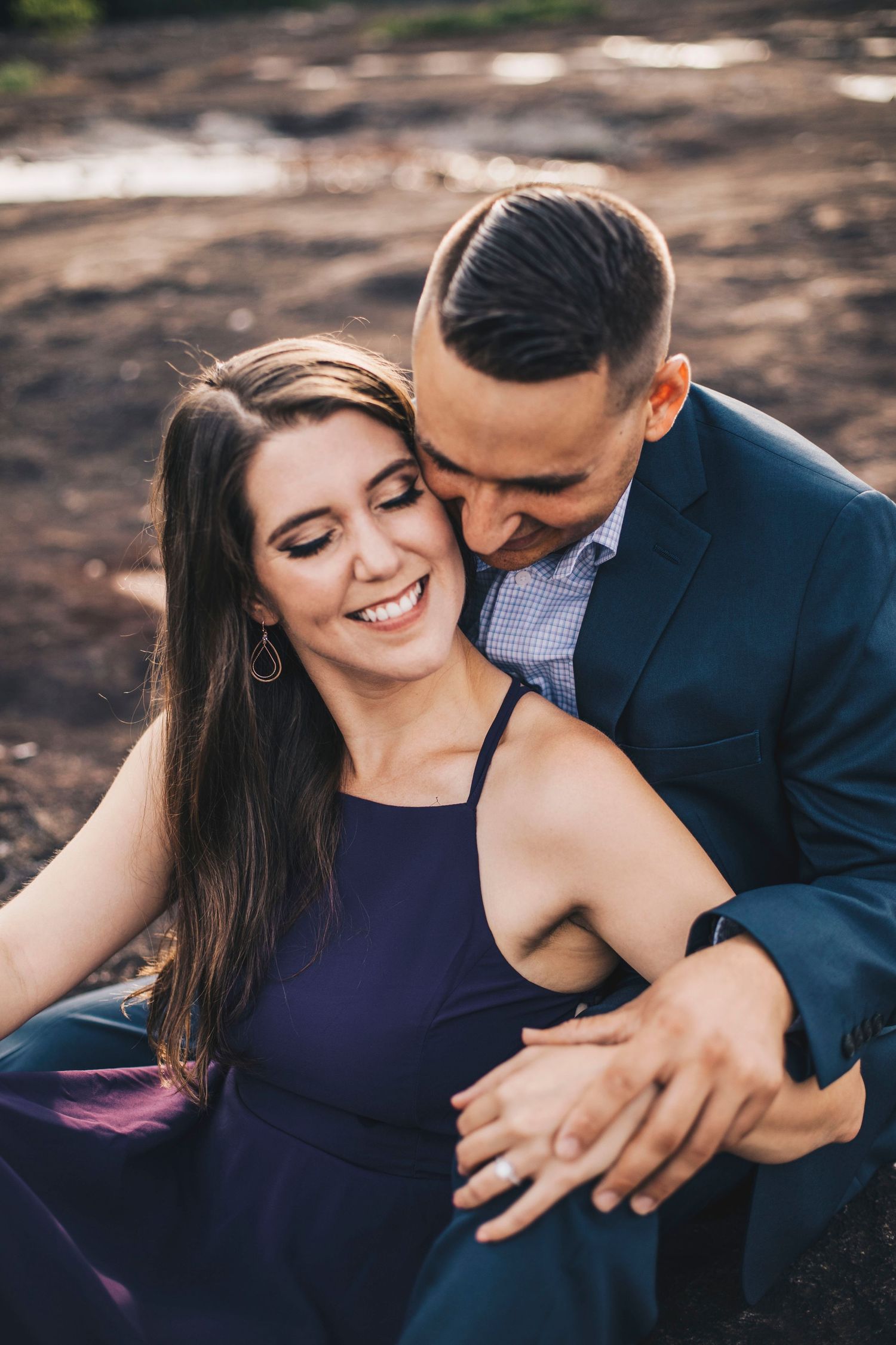 Romantic couple embracing outdoors, woman in purple dress smiling as man kisses her cheek.
