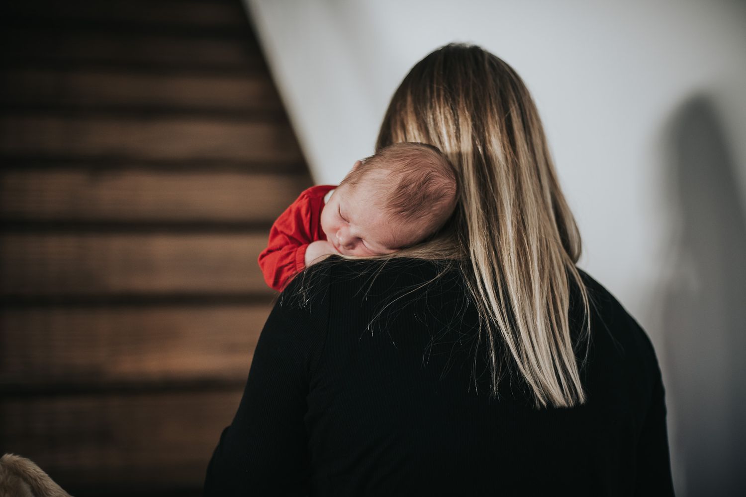 A sleeping baby rests on their parent's shoulder in front of a wooden staircase.