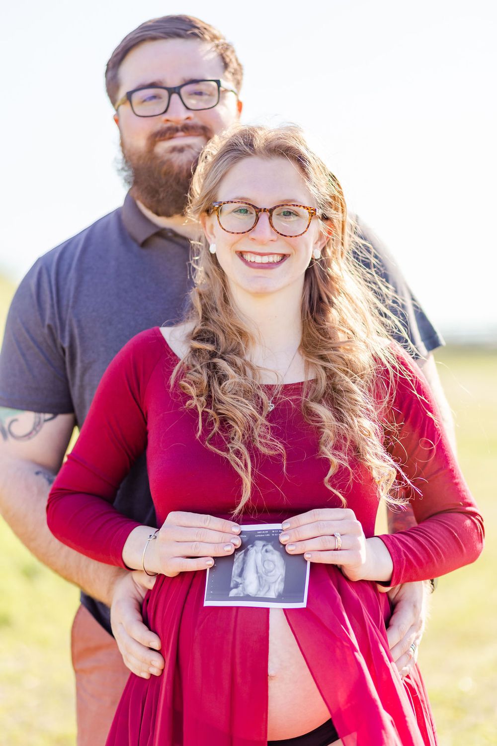A pregnancy announcement photo showing an ultrasound image held against a red dress outdoors.