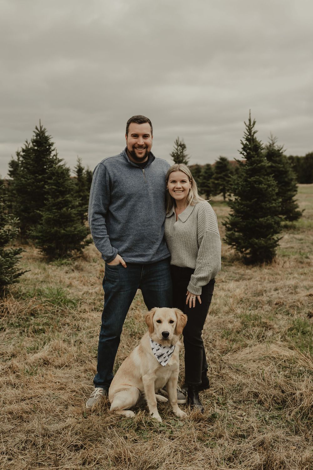 A couple and their Yellow Labrador dog pose together among evergreen trees on an overcast day.