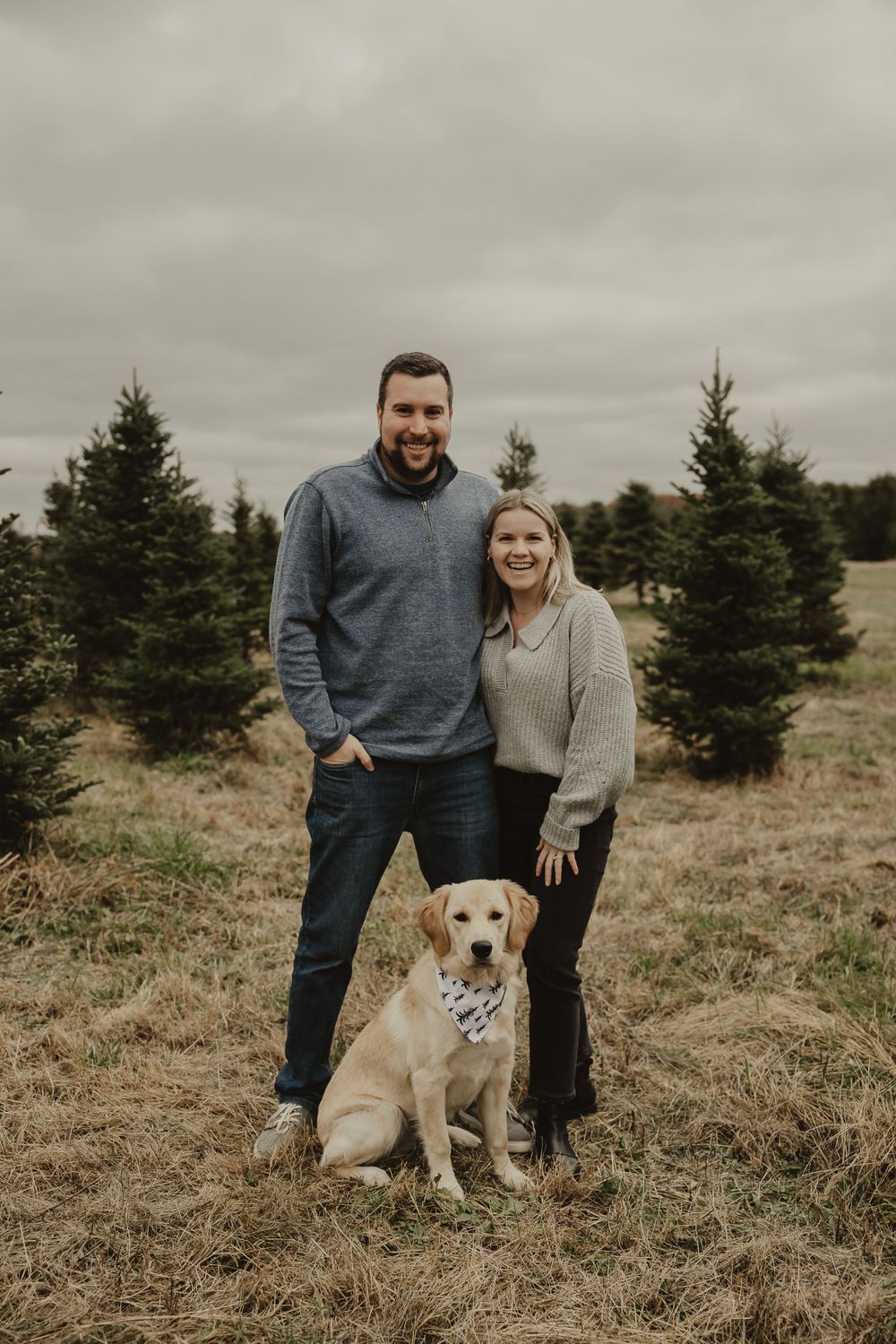 A couple with their Labrador Retriever standing between pine trees on a cloudy afternoon.