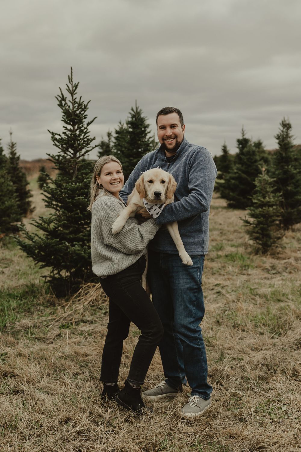 A couple holds their happy Yellow Lab puppy while standing in a Christmas tree field.