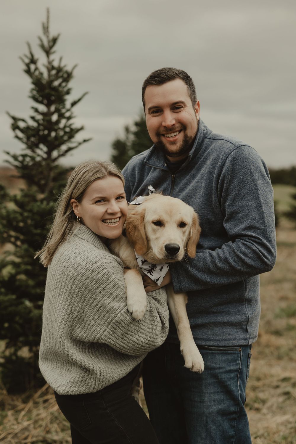 A couple smiles while holding their Yellow Lab puppy in a field of Christmas trees.