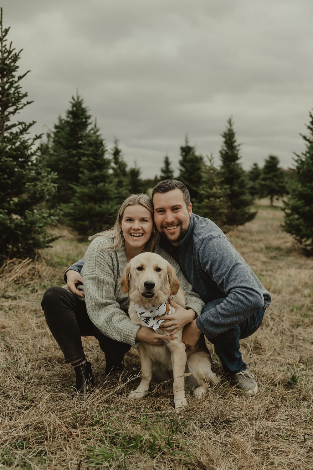 A couple and their golden retriever pose together among evergreen trees on a cloudy day in nature.