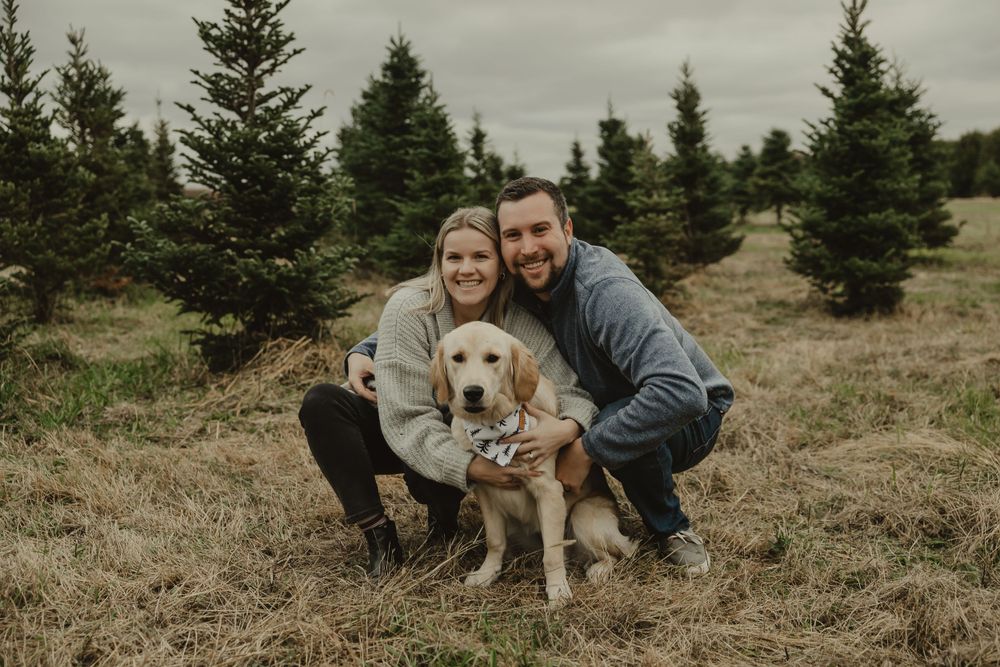 A couple crouches with their golden retriever in front of pine trees during an outdoor photo session.