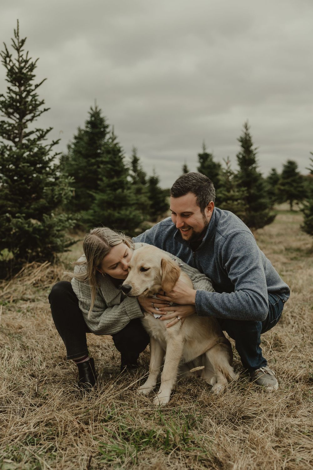A couple shares a tender moment with their golden retriever dog against a backdrop of evergreen trees.