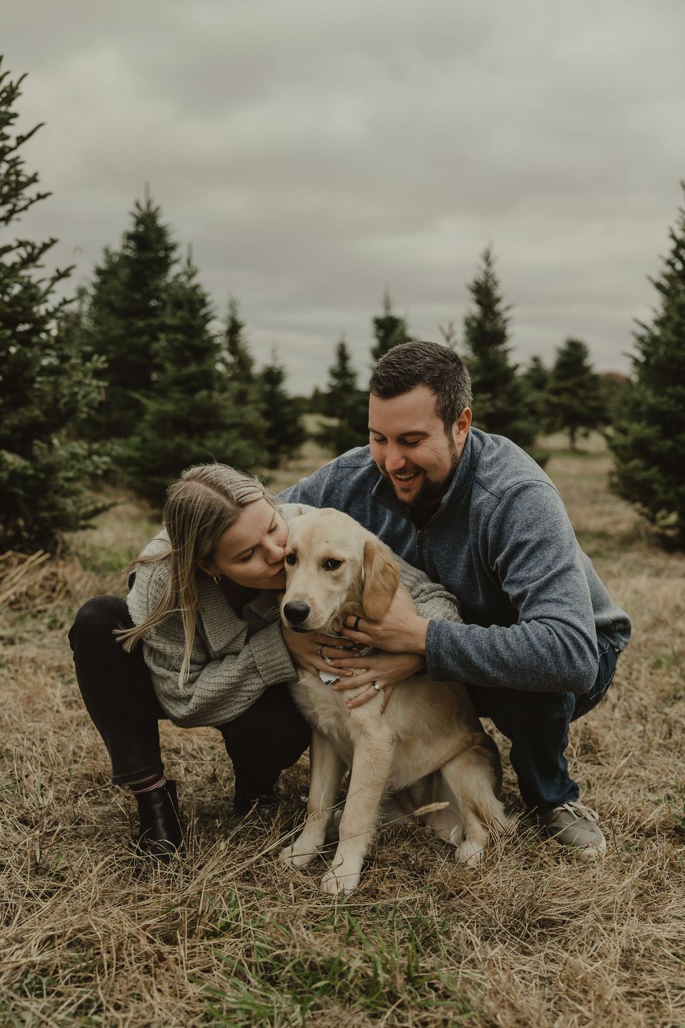 A couple embraces their golden retriever while crouching in a field surrounded by pine trees.