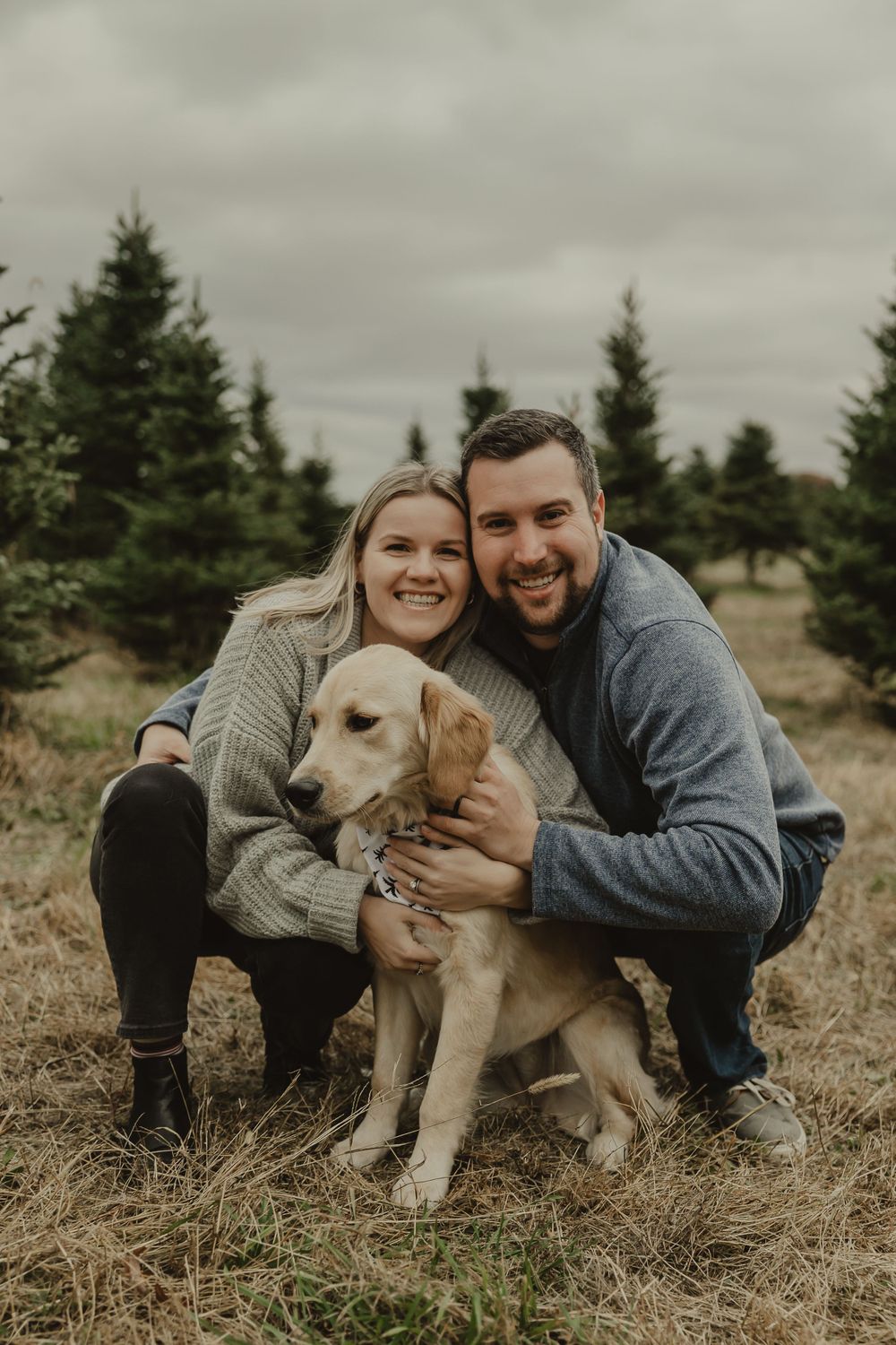 A couple poses with their golden retriever for a casual outdoor portrait among evergreen trees.