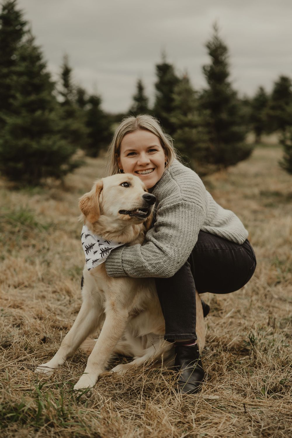 A person embraces their golden retriever dog while posing in a field with pine trees.