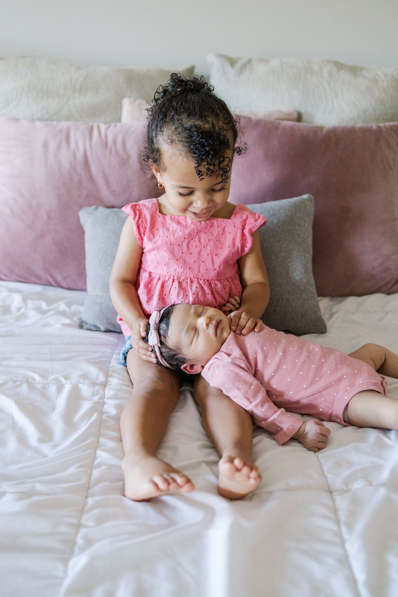 Two siblings share a tender moment on a bed with pink and white pillows.