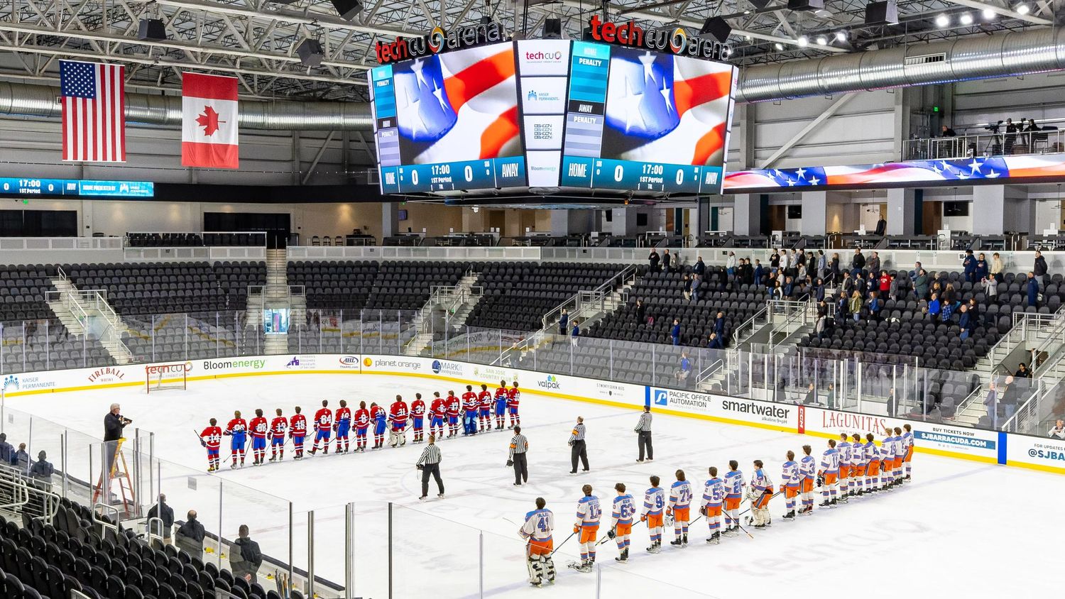 Hockey teams line up on ice rink with national flags displayed on arena screens.