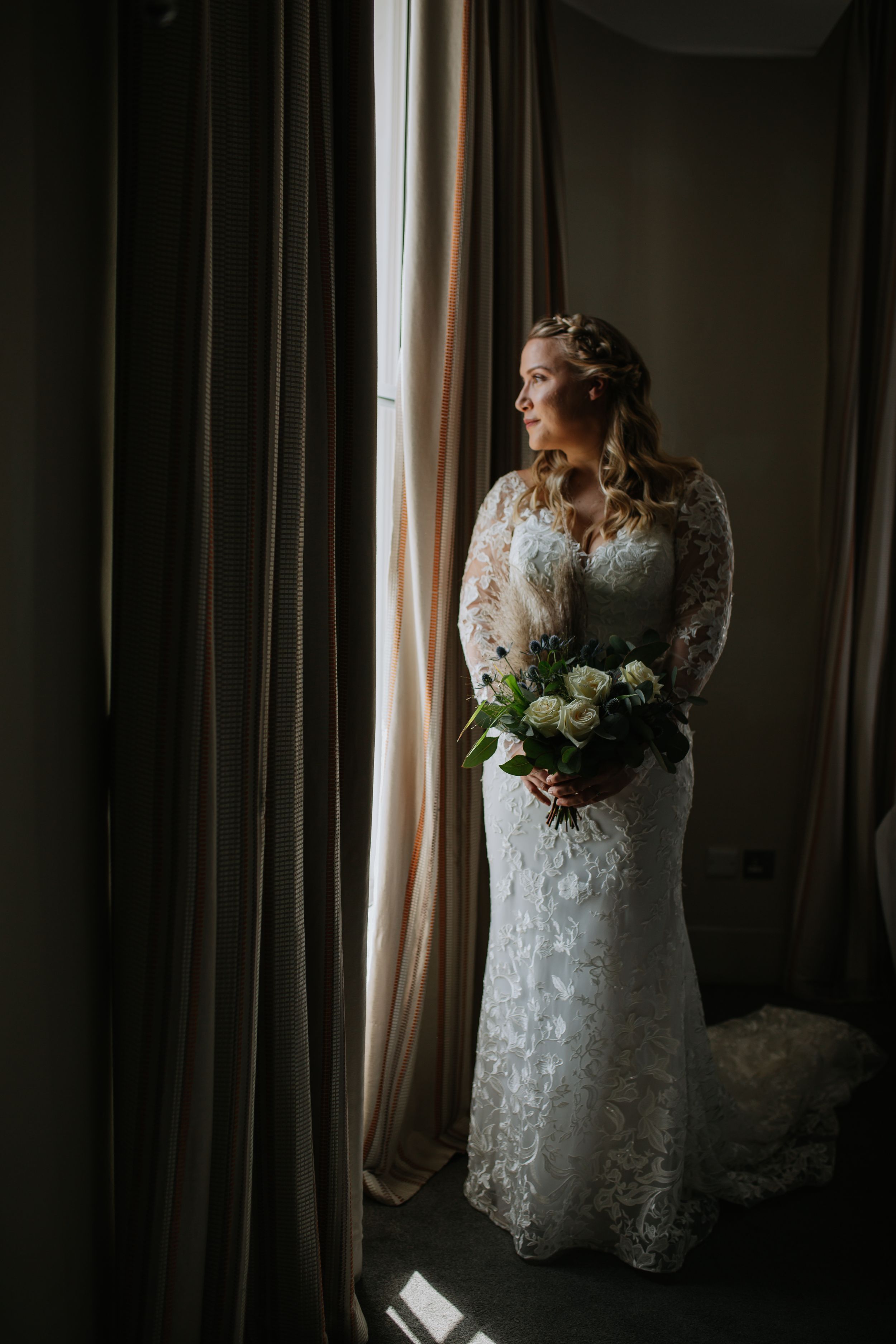 Bride in lace gown holds white bouquet while standing by window with flowing curtains.